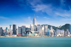 The modern Hong Kong skyline rises along the turquoise waters of Victoria Harbour under a bright blue sky.