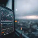 A computer monitor displays glowing financial charts next to a rainy window overlooking a blurred city skyline at dusk.