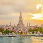 The grand spire of Wat Arun temple towers over the Chao Phraya River in Bangkok, Thailand, during a golden sunset sky.