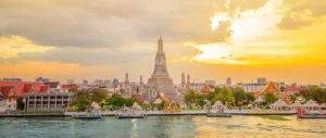 The grand spire of Wat Arun temple towers over the Chao Phraya River in Bangkok, Thailand, during a golden sunset sky.