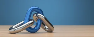A glossy blue chain link is interlocked with two silver metallic links on a wooden table against a solid blue background.