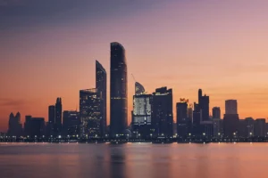 Illuminated modern skyscrapers line the Abu Dhabi waterfront against a vibrant orange and purple dusk sky.