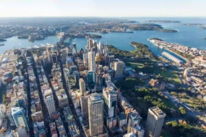 An aerial view of the Sydney skyline features dense downtown skyscrapers surrounding Sydney Tower next to the harbor.
