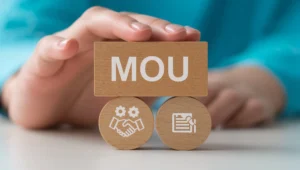 A hand holds a rectangular wooden block reading MOU positioned above circular blocks with handshake and document icons.