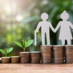 A paper cutout of an elderly couple stands on stacks of coins beside small growing saplings on a wooden surface.