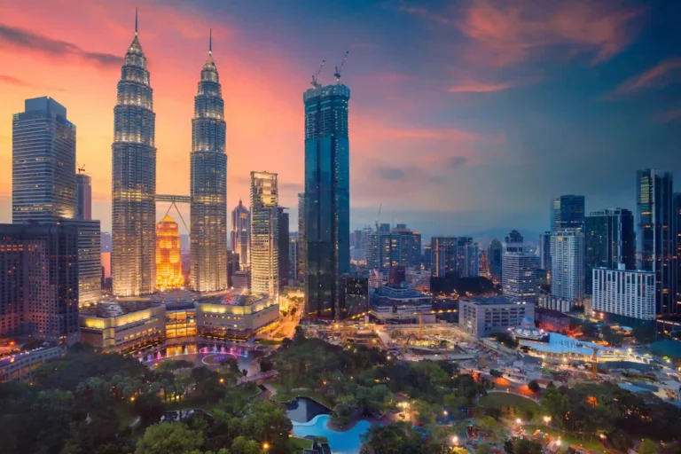 The illuminated Petronas Twin Towers stand tall over the Kuala Lumpur city skyline and park at sunset.