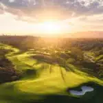 Sunlight streams over a sprawling green golf course with sand bunkers surrounded by rolling hills at sunset.