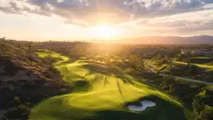 Sunlight streams over a sprawling green golf course with sand bunkers surrounded by rolling hills at sunset.