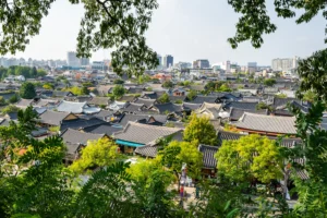 Tiled roofs of traditional Korean hanok houses fill Jeonju Village surrounded by leafy trees and modern city buildings.