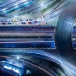 An aerial top-down view shows long light trails from traffic moving across a highway interchange at night.