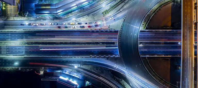 An aerial top-down view shows long light trails from traffic moving across a highway interchange at night.