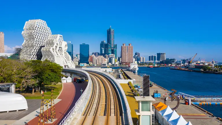 Light rail tracks curve along the Kaohsiung waterfront toward modern architectural landmarks and city skyscrapers.
