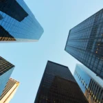 Modern glass skyscrapers reach upward toward a clear blue sky in a bustling city financial district.