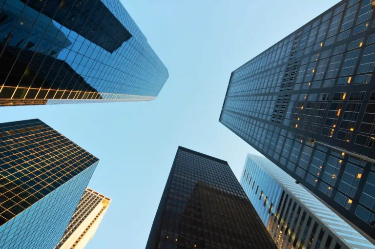 Modern glass skyscrapers reach upward toward a clear blue sky in a bustling city financial district.