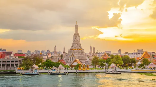 The grand spire of Wat Arun temple towers over the Chao Phraya River in Bangkok, Thailand, during a golden sunset sky.