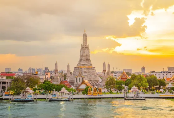 The grand spire of Wat Arun temple towers over the Chao Phraya River in Bangkok, Thailand, during a golden sunset sky.