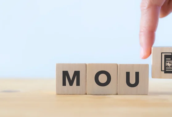 A hand places a wooden block with a handshake icon beside blocks spelling MOU on a light wood surface.